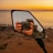 A camera is held up by a person capturing a reflection in a side mirror. The background features a scenic view of a body of water and a vibrant sunset sky. The scene is calm and picturesque, conveying a moment of photography during a sunset.