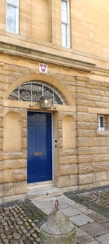 A stone building facade with detailed masonry work featuring a vibrant blue door centered under a stone arch. The arch is adorned with a decorative shield bearing a red symbol, and there is a large lamp mounted above the door. The ground is paved with stone and cobblestones, and a small stone bollard is visible in the foreground.