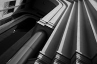 A black and white photograph highlighting a modern architectural structure. The image features a series of converging diagonal lines with deep shadows, emphasizing the geometric design and texture of the building.