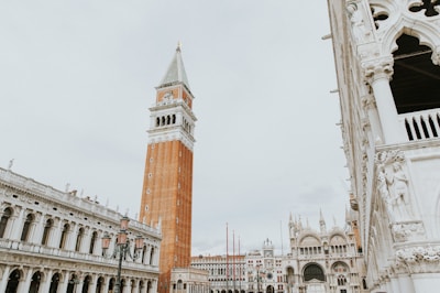 A tall brick bell tower stands prominently in the center, with ornate white stone buildings featuring arched windows and intricate sculptures surrounding it. The architectural style is classical, reflecting a historical European influence.