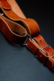 A close-up view of a brown leather belt with metal buckles, intricately placed on a dark background. The leather has a rich texture, with visible stitching and a sturdy appearance.