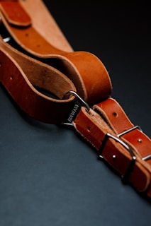 A close-up view of a brown leather belt with metal buckles, intricately placed on a dark background. The leather has a rich texture, with visible stitching and a sturdy appearance.