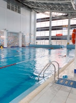 A spacious indoor swimming pool with clear blue water and lane dividers. The pool is surrounded by a tiled deck, with a ladder for access. The large windows on one side allow natural light to fill the area, and there is a large digital clock displayed on the wall. Life-saving equipment such as a lifebuoy is visible on the side. A pair of blue sandals is placed near the pool entrance.