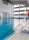 A spacious indoor swimming pool with clear blue water and lane dividers. The pool is surrounded by a tiled deck, with a ladder for access. The large windows on one side allow natural light to fill the area, and there is a large digital clock displayed on the wall. Life-saving equipment such as a lifebuoy is visible on the side. A pair of blue sandals is placed near the pool entrance.
