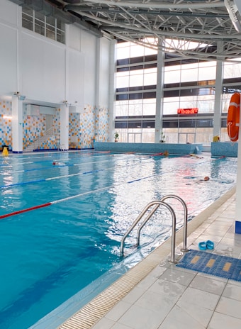 A spacious indoor swimming pool with clear blue water and lane dividers. The pool is surrounded by a tiled deck, with a ladder for access. The large windows on one side allow natural light to fill the area, and there is a large digital clock displayed on the wall. Life-saving equipment such as a lifebuoy is visible on the side. A pair of blue sandals is placed near the pool entrance.