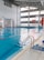 A spacious indoor swimming pool with clear blue water and lane dividers. The pool is surrounded by a tiled deck, with a ladder for access. The large windows on one side allow natural light to fill the area, and there is a large digital clock displayed on the wall. Life-saving equipment such as a lifebuoy is visible on the side. A pair of blue sandals is placed near the pool entrance.