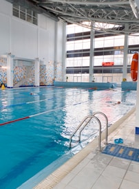 A spacious indoor swimming pool with clear blue water and lane dividers. The pool is surrounded by a tiled deck, with a ladder for access. The large windows on one side allow natural light to fill the area, and there is a large digital clock displayed on the wall. Life-saving equipment such as a lifebuoy is visible on the side. A pair of blue sandals is placed near the pool entrance.