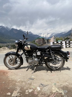 A classic motorcycle is parked on the side of a road with a mountainous landscape in the background. Snow-capped peaks are visible under a cloudy sky, and nearby buildings are partially seen, giving a rustic and adventurous vibe.