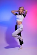 A woman with blonde hair stands in a dance pose with one leg raised and her arms touching her head. She wears a cropped, short-sleeved white shirt and black and white athletic pants in a minimalistic studio with gradient lighting transitioning from blue on the left to pink on the right.