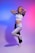 A woman with blonde hair stands in a dance pose with one leg raised and her arms touching her head. She wears a cropped, short-sleeved white shirt and black and white athletic pants in a minimalistic studio with gradient lighting transitioning from blue on the left to pink on the right.