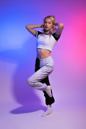 A woman with blonde hair stands in a dance pose with one leg raised and her arms touching her head. She wears a cropped, short-sleeved white shirt and black and white athletic pants in a minimalistic studio with gradient lighting transitioning from blue on the left to pink on the right.