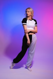 A person with long blonde hair stands in a poised, fashionable pose. They are wearing a black and white athletic outfit, consisting of a short-sleeved top and pants, set against a background featuring a gradient of blue to pink colors.