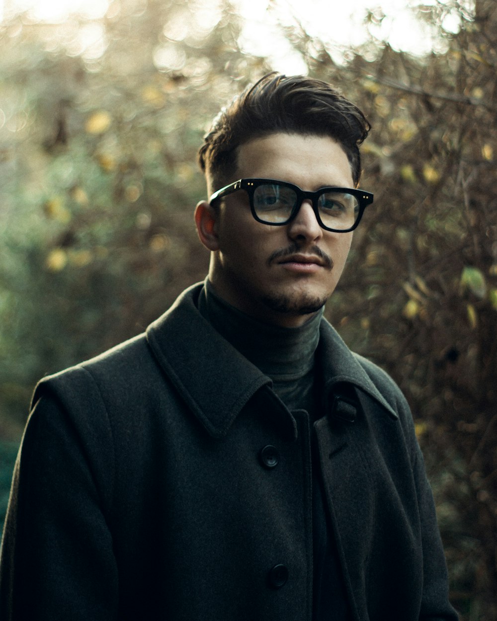 a man wearing glasses standing in front of a tree