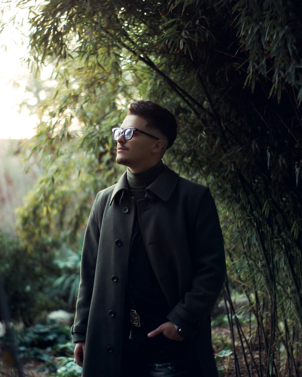 a man wearing a coat and sunglasses standing in a forest