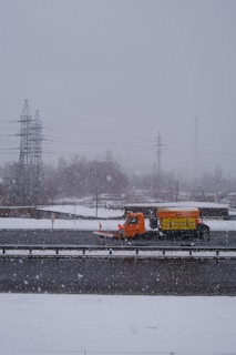 An orange snowplow truck is clearing snow from a highway during a snowfall. The scene is dominated by an overcast sky with snowflakes visibly falling. In the background, there are utility poles and power lines along with some bare trees partially obscured by the snow.