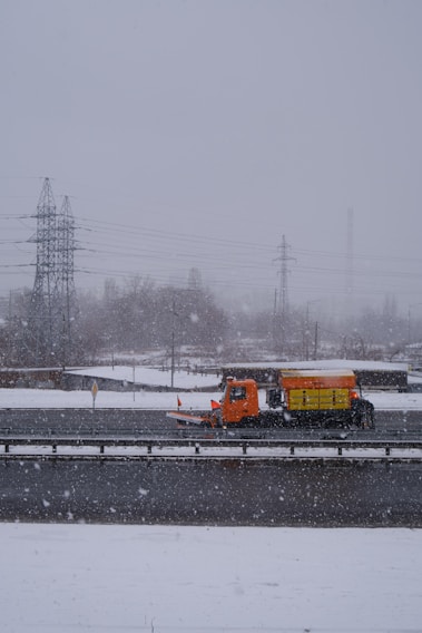 An orange snowplow truck is clearing snow from a highway during a snowfall. The scene is dominated by an overcast sky with snowflakes visibly falling. In the background, there are utility poles and power lines along with some bare trees partially obscured by the snow.