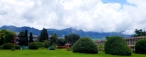 A lush green landscape with manicured bushes and trees is set against a backdrop of rolling mountains partially obscured by a layer of clouds. A large building with a modernist architectural style can be seen, featuring windows and a flat roof.