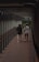 Two individuals walk side by side through a covered pedestrian walkway with wooden flooring and metal railings. The path is enclosed on both sides with glass and metal structural elements. The lighting inside is dim, accentuated by small overhead fixtures. Outside the walkway, greenery is visible.