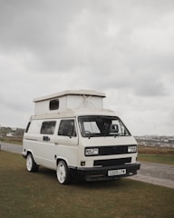 A white camper van with distinctive square headlights and a pop-up roof parked on a grassy area. Overcast skies create a muted background, and there is a road and additional landscape visible in the distance.