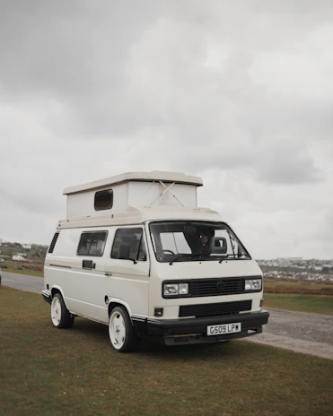 A white camper van with distinctive square headlights and a pop-up roof parked on a grassy area. Overcast skies create a muted background, and there is a road and additional landscape visible in the distance.
