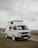 A white camper van with distinctive square headlights and a pop-up roof parked on a grassy area. Overcast skies create a muted background, and there is a road and additional landscape visible in the distance.