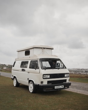 A white camper van with distinctive square headlights and a pop-up roof parked on a grassy area. Overcast skies create a muted background, and there is a road and additional landscape visible in the distance.