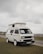 A white camper van with distinctive square headlights and a pop-up roof parked on a grassy area. Overcast skies create a muted background, and there is a road and additional landscape visible in the distance.