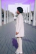 A person with curly hair, wearing a cream-colored coat and pants, stands on a boardwalk lined with modern, artistic pillars. The background features a large white structure with the text 'PORT VELL' and a purple sky. The setting appears urban and contemporary.
