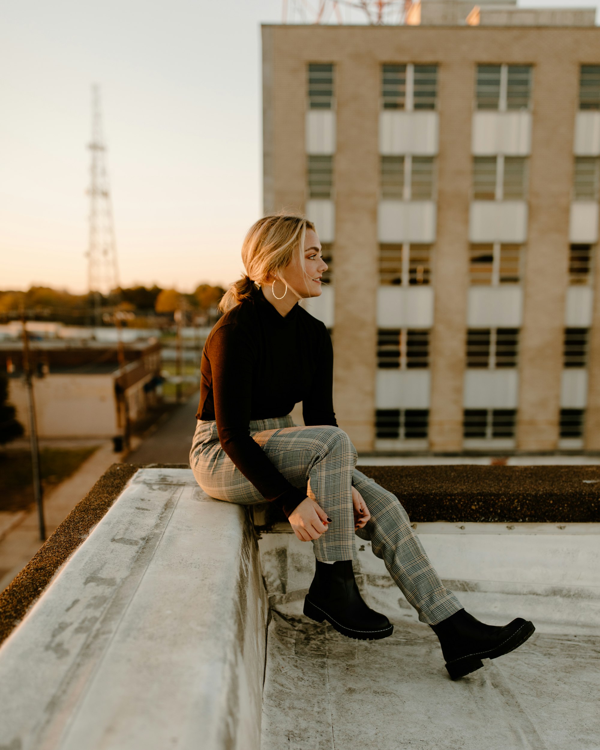 hip young woman in a burgundy turtleneck sweater sitting on corner rooftop building looking out at sunset