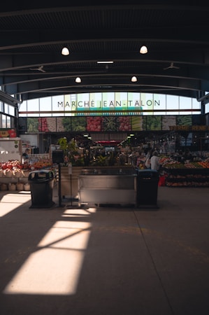 An indoor market scene with the name 'March&eacute; Jean-Talon' prominently displayed at the front, surrounded by images of fresh produce. Natural light streams through large windows, illuminating the produce stands and a few people shopping. The atmosphere is spacious, showcasing a variety of fruits and vegetables in an organized manner.