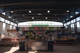A bustling indoor marketplace with high ceilings and abundant natural light. Stalls are filled with various fresh produce, including fruits and vegetables. The name 'Marché Jean-Talon' is prominently displayed above, and shoppers can be seen browsing and purchasing items.