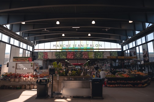A bustling indoor marketplace with high ceilings and abundant natural light. Stalls are filled with various fresh produce, including fruits and vegetables. The name 'Marché Jean-Talon' is prominently displayed above, and shoppers can be seen browsing and purchasing items.