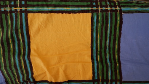 A fabric pattern featuring large blocks of color, including a prominent yellow square and a blue section. The pattern is bordered and intersected by black, green, and turquoise stripes, creating a grid-like design.