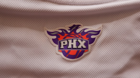 A logo set against a textured white fabric background, featuring a stylized phoenix above a basketball. The letters 'PHX' are prominently displayed in a bold font at the center.