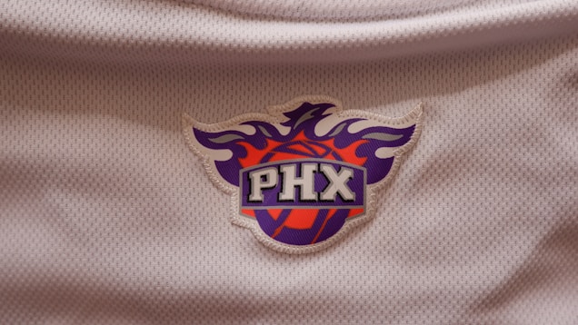A logo set against a textured white fabric background, featuring a stylized phoenix above a basketball. The letters 'PHX' are prominently displayed in a bold font at the center.