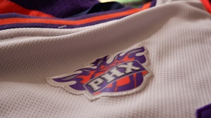A close-up view of a sports jersey featuring a logo with the letters 'PHX' on a basketball design. The jersey is predominantly white with orange and purple accents.