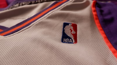 A close-up of a sports jersey made of mesh fabric features the NBA logo prominently. The jersey has an orange and purple trim on a white background.