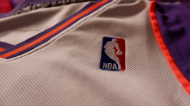 A close-up of a sports jersey made of mesh fabric features the NBA logo prominently. The jersey has an orange and purple trim on a white background.