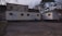 Two mobile trailers are situated in front of a brick building with graffiti. The area is surrounded by leafless trees, and there is a traffic cone and some trash bins next to the trailers. The ground is paved, and the scene has a somewhat neglected, industrial feel.