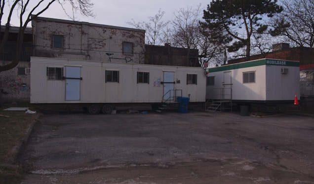 Two mobile trailers are situated in front of a brick building with graffiti. The area is surrounded by leafless trees, and there is a traffic cone and some trash bins next to the trailers. The ground is paved, and the scene has a somewhat neglected, industrial feel.