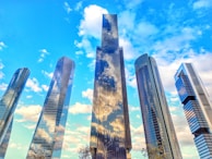 A group of modern skyscrapers with reflective glass surfaces stand against a vibrant blue sky speckled with fluffy white clouds. The buildings vary in height and feature sleek, curved, and angular designs, creating a sense of architectural innovation.