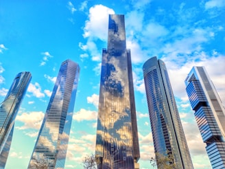 A group of modern skyscrapers with reflective glass surfaces stand against a vibrant blue sky speckled with fluffy white clouds. The buildings vary in height and feature sleek, curved, and angular designs, creating a sense of architectural innovation.