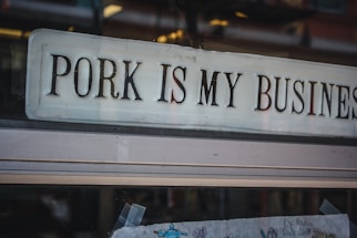 A storefront sign displaying the phrase 'PORK IS MY BUSINESS' in bold, uppercase letters. The sign is placed in a glass window, and below it are some drawings, possibly made by children, with visible tape holding them in place. The image captures a reflection of the street in the glass.