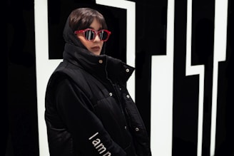 A person wearing a black hooded jacket and red sunglasses stands against a bold, geometric black and white background. The jacket has a high collar and the word 'Iam' is visible on the sleeve, adding a touch of identity to the attire.