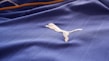 A close-up of a textured blue fabric with a white embroidered logo of a leaping animal, likely representing a brand. The fabric appears to be part of a sports jersey, with fine linear stitching visible.