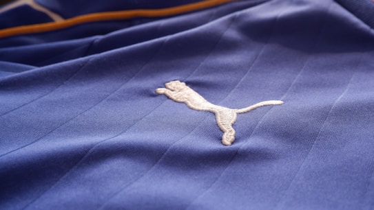 A close-up of a textured blue fabric with a white embroidered logo of a leaping animal, likely representing a brand. The fabric appears to be part of a sports jersey, with fine linear stitching visible.