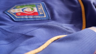 A blue fabric with a badge displaying the Italian flag colors and an emblem featuring stars is seen. The garment has gold trim along the seams.