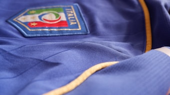 A blue fabric with a badge displaying the Italian flag colors and an emblem featuring stars is seen. The garment has gold trim along the seams.