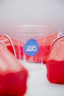 A clear plastic cup with a blue sticker displaying the letters 'ec' is surrounded by several red solo cups. The red cups are haphazardly arranged and provide a stark contrast against the clear cup, which is centrally positioned and highlighted.