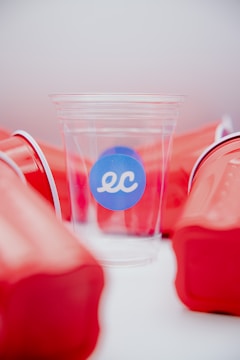 A clear plastic cup with a blue sticker displaying the letters 'ec' is surrounded by several red solo cups. The red cups are haphazardly arranged and provide a stark contrast against the clear cup, which is centrally positioned and highlighted.